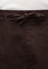 Κοντινή λήψη σκούρων καφέ παντελονιών με δέσιμο υφασμάτινου κορδονιού στη μέση, φορεμένα πάνω από λευκό ριμπ πουκάμισο.