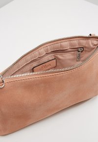 Różowy zamszowy kosmetyczka z zamkiem błyskawicznym, wyposażona w wewnętrzną kieszeń i małą metkę z logo. Gładka faktura i minimalistyczny design.