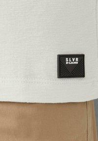 Witte, textuurstof met een zwart rechthoekig label met opstaande opdruk; het label leest "SLVR BY S.OLIVER" in witte letters. Bruine stof eronder.