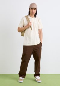 Polo beige con maniche corte, pantaloni marroni, cappellino beige e sneakers bianche. Il modello porta uno zaino kaki su uno sfondo neutro.