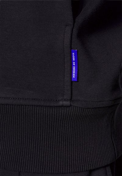 O sweatshirt preto apresenta um acabamento canelado na barra e nos punhos, com uma etiqueta retangular azul rotulada "TIGER OF SWEDEN" perto da costura. Tecido suave e texturizado.