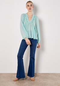 Helles blaues, langärmeliges Bluse mit V-Ausschnitt und dekorativen Knöpfen; kombiniert mit dunklen, eingestellten Jeans und beigen Sandalen mit Riemchen.