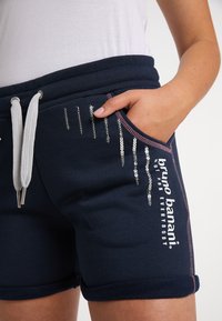Marineblauwe shorts met pailletten details, elastische tailleband met trekkoord, roze stikselaccenten en zijzakken. Merklogo zichtbaar.