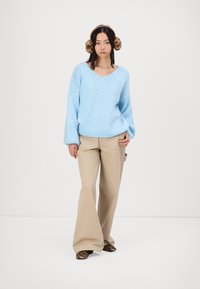 Lichtblauwe gebreide trui met V-hals, gecombineerd met beige brede broek en slippers met luipaardprint; model draagt bruine nepbont oorwarmers.