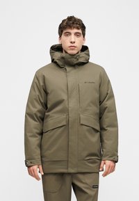 Άνδρας που φοράει χακί πράσινο μπουφάν Columbia με κουκούλα και αντίστοιχο παντελόνι, κοιτάζοντας μπροστά σε απλό ανοιχτό φόντο.