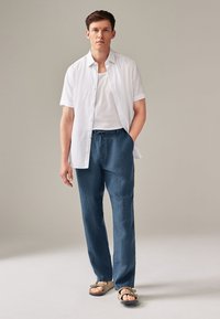 Camicia bianca leggera a maniche corte, canotta grigia, pantaloni in lino blu e sandali marroni, con una vestibilità rilassata e vita con cordino.
