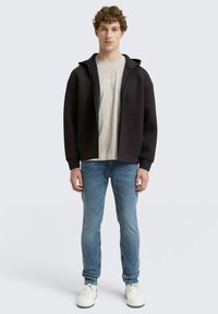 Fekete kapucnis sweatshirt, elöl cipzárral és két zsebbel, világosszürke póló felett viselve. Világoskék farmernadrággal és fehér tornacipővel párosítva.