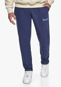 Marineblå sweatpants i blødt materiale med sidelommer og detaljer med hvidt logo på låret; parret med hvide sneakers.