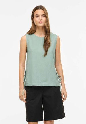 Femme portant un top sans manches vert clair avec des liens sur les côtés et un short noir ample, debout sur un fond blanc.