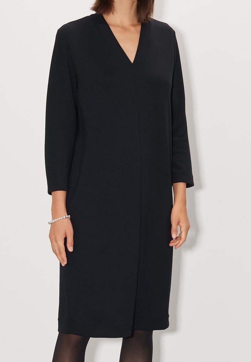 Černé šaty ke kolenům s výstřihem do V, dlouhými rukávy a rovnou siluetou. Vyrobené z texturované tkaniny bez švů a viditelného kování.