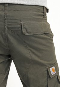 Cargo spodnie w oliwkowej zieleni z teksturowanej tkaniny, podwójnymi przeszyciami i boczną kieszenią z widoczną naszywką marki.