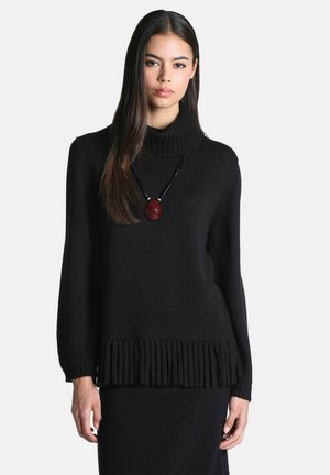 Femme portant un pull à col roulé côtelé noir et une jupe, avec de longs cheveux raides et un pendentif rouge sur un cordon noir.