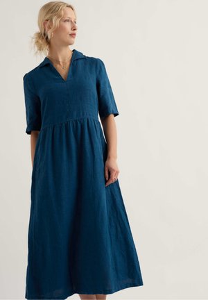 Szőke nő kék lenvászon midi ruhát visel, amelynek könyékig érő ujjai és gallérja van, balra néz egy egyszínű háttér előtt.