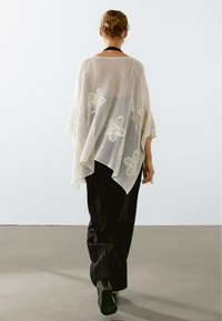 Kobieta z włosami związanymi w kok, ubrana w białą koronkową chustę z kwiatowymi wzorami, czarne spodnie i czarne buty na słupku, idąca w stronę przeciwną.