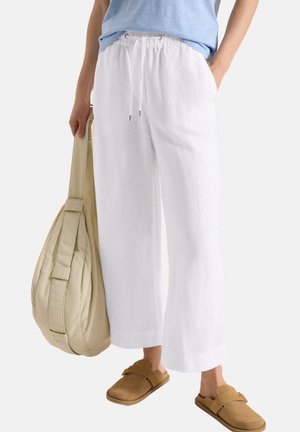 Osoba nosząca luźne białe spodnie i beżowe buty wsuwane, trzyma dużą beżową torbę z paskiem.