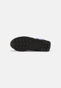 Semelle en caoutchouc noir d'une chaussure de trail avec des motifs de crampons triangulaires profonds pour une adhérence renforcée sur terrain accidenté.