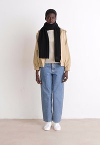 Giacca a vento beige, sciarpa nera a coste, felpa color crema, jeans larghi azzurri chiari e sneaker bianche. Look semplice e casual da streetwear.