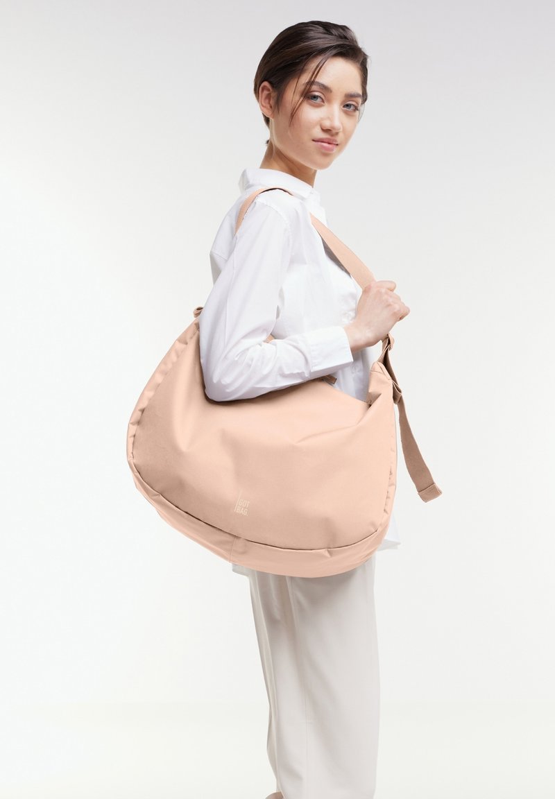 Borsa a spalla in tessuto rosa chiaro con forma arrotondata, tracolla regolabile e un piccolo dettaglio del logo. Texture liscia e design minimalista.