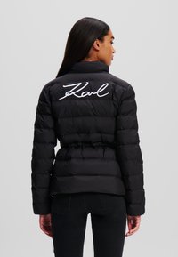 Schwarze Daunenjacke mit einem taillierten Gürtel, hoher Kragen und weißem gesticktem "Karl"-Logo auf dem Rücken. Glatte Textur, gesteppter Look.