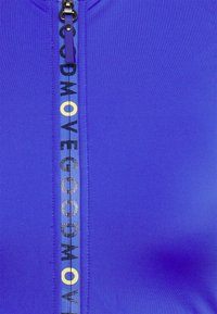 Top de desporto roxo com fecho de correr, apresentando um logótipo vertical em preto e amarelo ao longo da linha do fecho. Textura suave e forma ajustada.