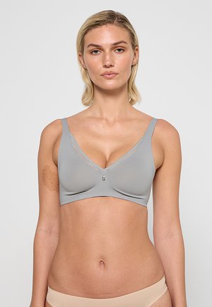 Woman with short blonde hair wearing a light gray seamless bra and nude underwear, standing against a plain light background.