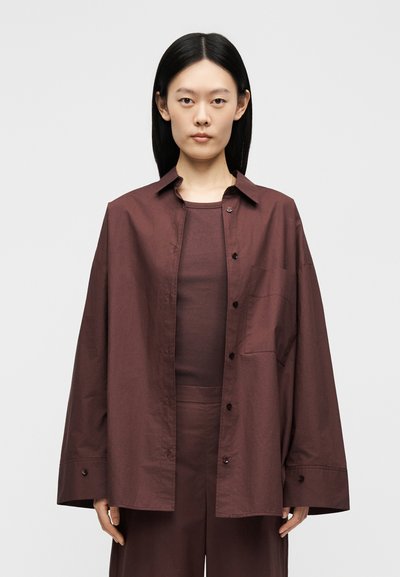 Jeune femme aux cheveux noirs lisses portant une chemise boutonnée oversize marron foncé sur un top et un pantalon assortis, debout devant un fond uni.