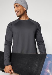 Dunkelgraues Langarmshirt mit Raglanärmeln, kombiniert mit einer grauen Strickmütze, und hält ein schwarzes Snowboard mit einem bunten Akzentrand.