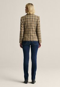 Nainen, jolla on lyhyet vaaleat hiukset, pukeutuneena vaaleanbeigeen ruudulliseen bleiseriin, tummansinisiin farkkuihin ja mustiin korkokenkiin, seisoo selkä kameraan päin.