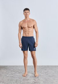 Tengeri kék úszónadrág elasztikus derékpánttal és oldalzsebekkel, bal lábon egy kis fehér logóval. Simább, könnyű anyagból.