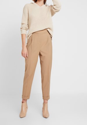 Femme portant un pull en maille beige, un pantalon beige taille haute avec revers roulés, et des bottines beige assorties, debout devant un fond uni.