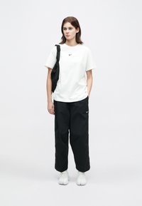 Egy nő áll fehér Nike pólóban, laza fekete Nike nadrágban, fehér sportcipőben, és egy fekete válltáskával egy fehér háttér előtt.