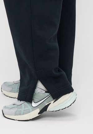 Pantalon noir avec fentes latérales reposant sur des pieds chaussés de baskets Nike bleu clair et gris avec des chaussettes blanches sur un fond uni.
