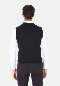 Gilet nero in maglia con design aderente, collo e orlo a costine, indossato sopra una camicia bianca a maniche lunghe, visto da dietro.