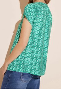 Zielona bluzka z krótkim rękawem o geometrycznym wzorze liści. Wykonana z miękkiego materiału, ma luźny krój, okrągły dekolt oraz detal w postaci plisy na plecach.
