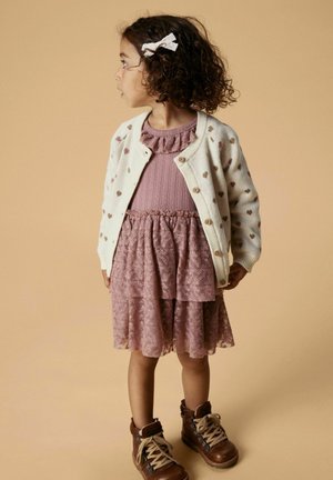 Cremefarbener Cardigan mit Herzmustern, übereinander liegendes rosa Spitzenkleid, braune Schnürstiefel und eine weiße Schleife im lockigen Haar.