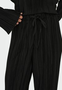 Pantalons plissés noirs avec taille à cordon de serrage et larges manches. Comprend des nervures verticales et une texture douce. Convient pour un usage décontracté.