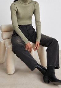 Prążkowana, z wysokim kołnierzem, długo rękawowa bluzka w oliwkowym kolorze; zestawiona z czarnymi jeansami z wysokim stanem; model nosi czarne botki na kostkę; minimalistyczne otoczenie.