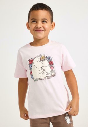 Smilende ung dreng iført en lyserød T-shirt og brune shorts, begge med tegneseriefigurer og blomsterdesign.