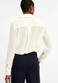 Weißes Blusenoberteil mit Rückeneinsatz, langen Ärmeln und geknöpften Manschetten. Kombiniert mit navyblauen, gestreiften Hosen, die glatte Texturen und ein einfaches Design zeigen.