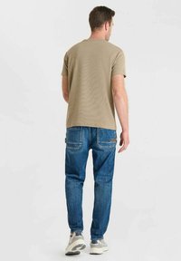 Camiseta de manga corta en color caqui combinada con jeans de mezclilla azules. Los jeans tienen costuras visibles y dos bolsillos traseros. Zapatillas deportivas blancas.
