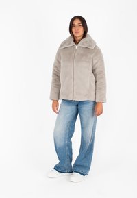 Giacca in pelliccia artificiale grigia chiara con un ampio colletto, chiusura frontale con zip e tasche laterali, abbinata a jeans in denim azzurri larghi e sneakers bianche.