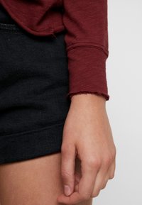 Bordowy top z długim rękawem o teksturowanej tkaninie, z surowym wykończeniem. W połączeniu z ciemnoniebieską spódnicą dżinsową, na której widać rękę odpoczywającą na udzie.