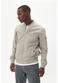 Hellgraue Bomberjacke mit Reißverschluss, gerippten Bündchen und Saum. Verfügt über Seitentaschen mit schwarzen Druckknöpfen und eine subtile, strukturierte Oberfläche.