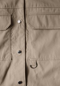 Beige tygplagg med två bröstfickor, glänsande silverknappar och en metall D-ring på framsidan för att fästa föremål. Slät textur.