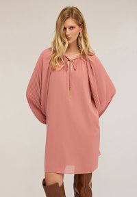 Donna che indossa un abito rosa largo con maniche a farfalla plissettate, scollo a cravatta, dettaglio con frange dorate e stivali di pelle marrone al ginocchio.