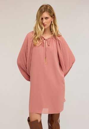 Vrouw in een losse roze jurk met geplooide vleermuismouwen, striknek, gouden franjes en kniehoge bruinleren laarzen.