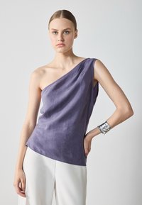 Violet vienos peties palaidinė, pagaminta iš minkštos, tekstūruotos medžiagos. Modelis ją dėvi su baltais kelnėmis ir sidabriniu apyrankiu.