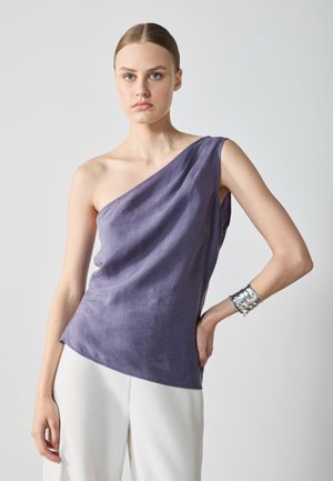 Purple one-shoulder top made from soft, textured fabric. Model wears it with white pants and a silver cuff bracelet.