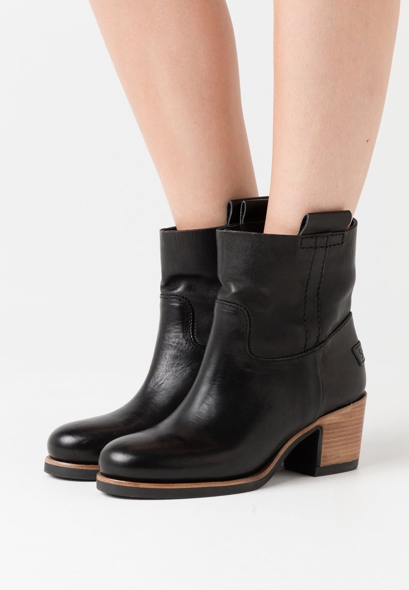 Bottines en cuir noir avec un bout rond et un talon en bois empilé. Présente des coutures détaillées et des tirettes pour un enfilage facile.