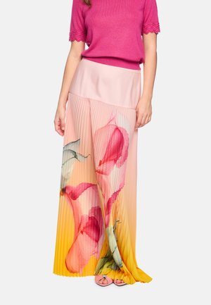 Woman wearing a pink top and a long pleated skirt with large floral patterns in pink, yellow, and green shades.